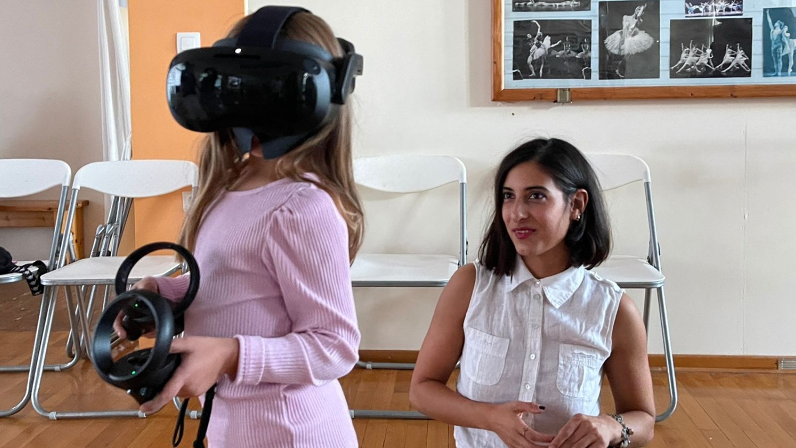 A young elementary school girl wearing a VR headset participates in a dance class. She stands with her arms gracefully extended, mimicking dance movements, while other children and an instructor practice in the background. The room is bright and spacious, with large mirrors reflecting the scene. The girl’s expression conveys focus and excitement as she immerses herself in the virtual experience.