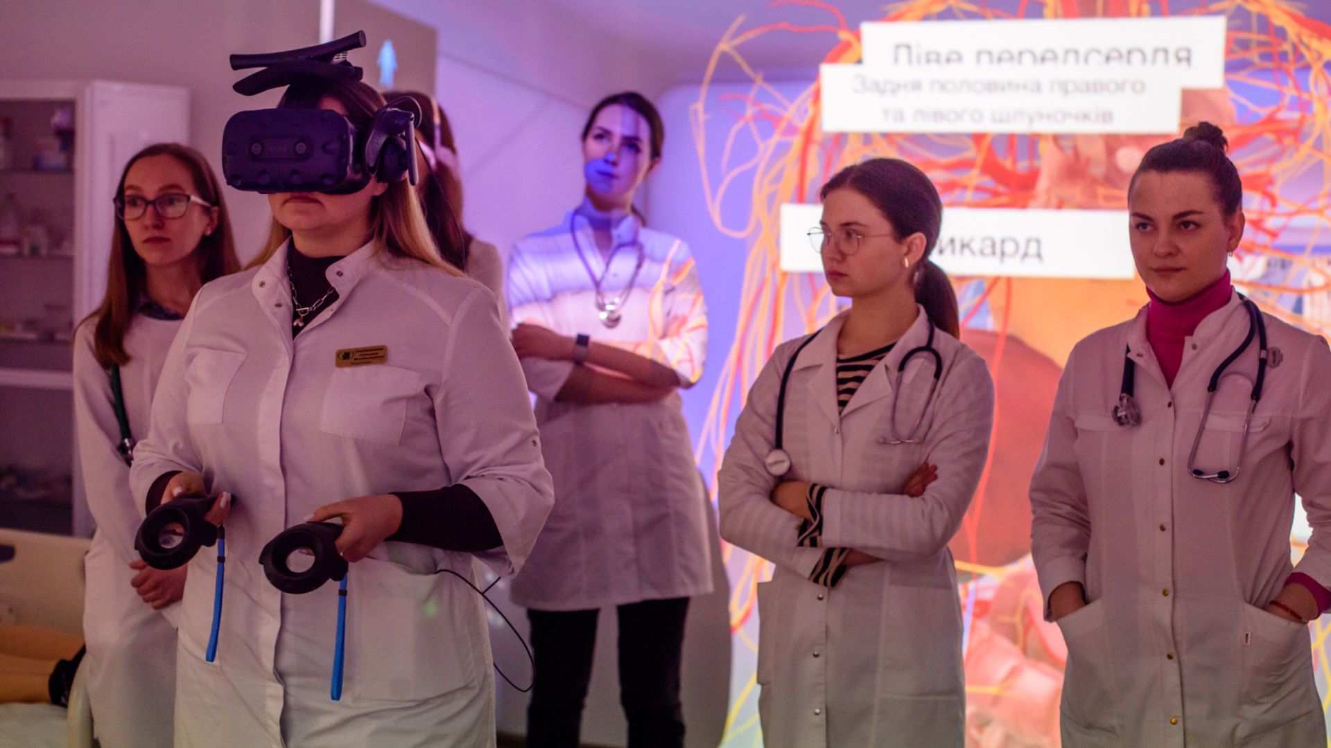 A group of medical students at Zaporizhzhia State Medical and Pharmaceutical University (ZSMPhU) participate in an immersive anatomy lesson. One student wears a VR headset and holds motion controllers, engaging with 3D anatomical content, while others observe attentively. The background features a projected anatomical diagram in Ukrainian, enhancing the interactive learning environment. All students are dressed in white lab coats and appear focused and engaged.